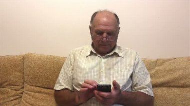 Yaşlı bir adam akıllı telefonuyla haber yayını okuyor, evdeki kanepede oturuyor. Gözlüklü ve beyaz gömlekli çekici yaşlı bir adam akıllı telefonunda haber okuyor, evdeki bir kanepede oturuyor..