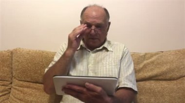 Yaşlı bir adam evdeki bir kanepede otururken tablet üzerinde çalışıyor. Gözlüklü ve beyaz gömlekli çekici yaşlı bir adam, evdeki bir kanepede oturup tablet bilgisayardan haber yayını okuyor..