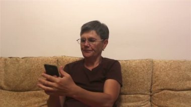 Evde bir kanepede otururken akıllı telefondan haber okuyan çekici yetişkin bir kadın. İnternette sörf yapmak için akıllı telefonunu kullanan bir kadın evdeki bir kanepede oturuyor..