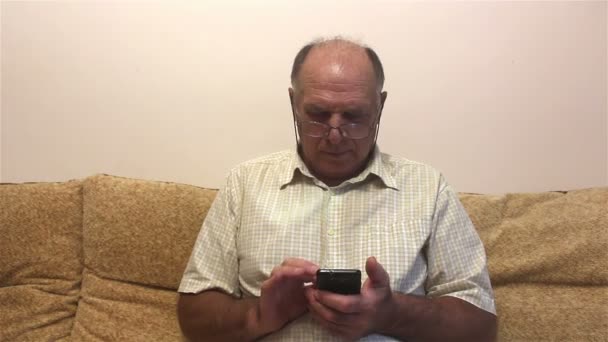 老人は自宅のソファに座って スマートフォンのニュースフィードを読み取ります 眼鏡と白いシャツを着た魅力的な高齢者は 自宅のソファに座って スマートフォンのニュースフィードを読んでいます — ストック動画