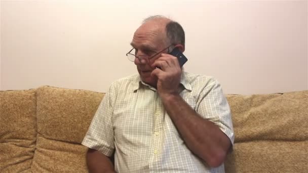 一位老人坐在沙发上 一边用智能手机打电话 一位戴眼镜 身穿白衬衫的漂亮老人一边坐在沙发上一边用手机聊天 一边坐在沙发上 — 图库视频影像