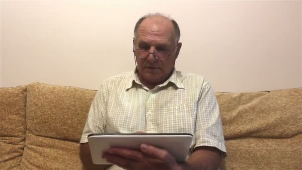 高齢者の男性は 自宅でソファに座っている間 タブレットPc上で動作します メガネと白いシャツを持つ魅力的な高齢者は タブレットPc上のニュースフィードを読んでいます 自宅でソファに座って — ストック動画