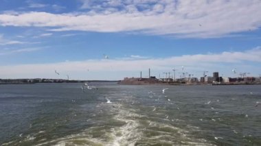 Güneşli havada gemiyi takip eden dalgaların görüntüsü. Martılar geminin arkasında uçar. Geminin kıç tarafından kıyı ve okyanus manzarası.