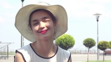 Yakın portre Beyaz şapkalı güzel bir genç kız kameraya bakar ve gülümser. Beyaz şapkalı genç ve çekici bir kız, bakışlarını gökyüzüne doğru kaldırıyor ve güneş ışınlarından gözlerini kısıyor..