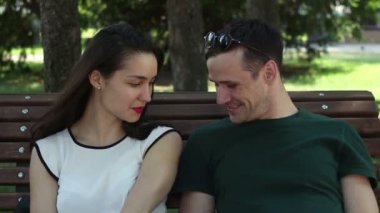 Beyaz tişörtlü çekici bir kız ve güneş gözlüklü ve yeşil tişörtlü genç yakışıklı bir adam parkta bir bankta otururken konuşuyor ve gülümsüyor..