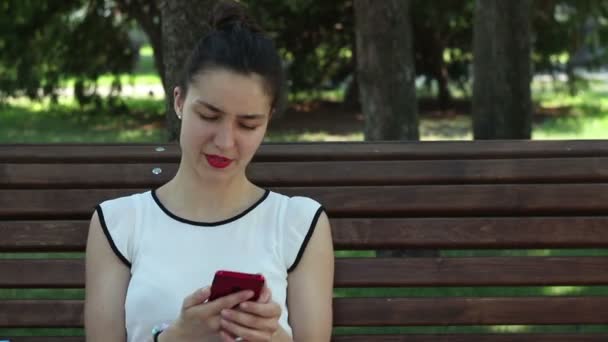 一位穿着白色T恤的年轻漂亮姑娘坐在公园的长椅上 一边用智能手机在社交网络上聊天 — 图库视频影像