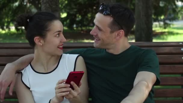 一位年轻漂亮的姑娘和她英俊的男朋友坐在公园的长椅上 一边用智能手机观看社交网络上的新闻 他们面带微笑地积极讨论新闻 — 图库视频影像