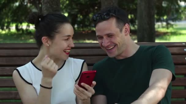 在一个阳光明媚的日子里 年轻的情侣们坐在公园的长椅上 正在积极讨论一款新的智能手机 他们面带微笑 互相拥抱 — 图库视频影像