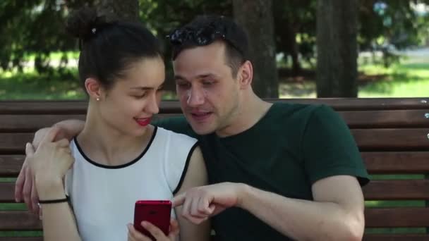 在一个阳光明媚的日子里 年轻的情侣们坐在公园的长椅上 正在积极讨论一款新的智能手机 他们面带微笑 互相拥抱 — 图库视频影像