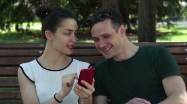 Güneşli bir günde seven çift, genç aktif olarak yeni bir akıllı telefon parkta bir bankta otururken tartışıyor. Gülümseyen ve birbirlerine sarılma.