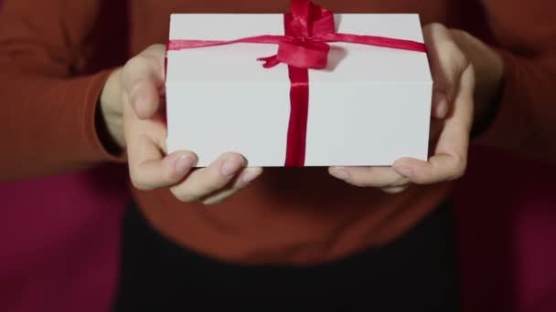 Hermosa chica sostiene un regalo en una caja blanca, con una cinta roja. Hermosas manos de niña sostienen un regalo de Año Nuevo, en una caja blanca con una cinta roja. Regalo para las vacaciones . — Vídeos de Stock