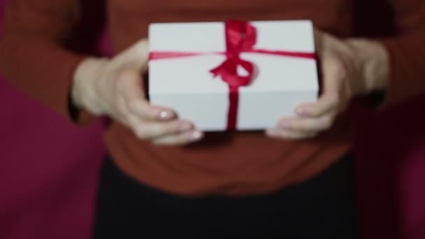 Menina bonita segura um presente em uma caixa branca, com uma fita vermelha. As mãos de menina bonitas mantêm um presente de Ano Novo, em uma caixa branca com uma fita vermelha. Presente para as férias . — Vídeo de Stock