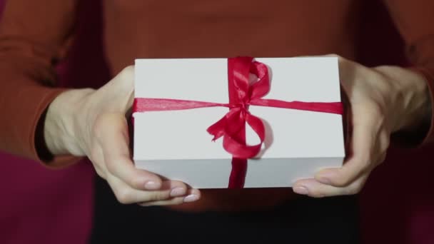 Hermosa chica sostiene un regalo en una caja blanca, con una cinta roja. Hermosas manos de niña sostienen un regalo de Año Nuevo, en una caja blanca con una cinta roja. Regalo para las vacaciones . — Vídeos de Stock