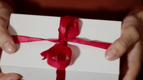 Hermosa chica abre su regalo. Hermosas manos de una chica abren un regalo de Año Nuevo, desatando una cinta roja. Regalo para las vacaciones. Regalo en una caja blanca con cinta roja . — Vídeo de stock