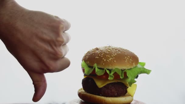 La mano masculina muestra el pulgar hacia abajo sobre un fondo de hamburguesa jugosa fresca. Hamburguesa grande y jugosa con chuleta de res, verduras frescas y queso derretido. Comida rápida, comida rica en calorías . — Vídeos de Stock