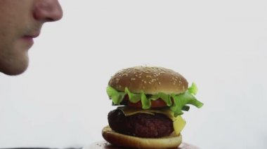 Young handsome guy is trying to bite a big juicy burger. Fast food, high-calorie food. Big juicy burger with beef cutlet, fresh vegetables and cream cheese. Burger close-up on a white background.