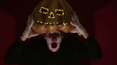 Scary man in the makeup of a clown holds a pumpkin for Halloween and puts it on his head.Terrible clown puts on a mask in the shape of a pumpkin on his head. Terrible clown holds in his hands a Jack-O