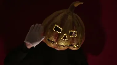 Scary man with a pumpkin head and luminous eyes looks at the camera. Man with a pumpkin head scares raising his hands up. Halloween.