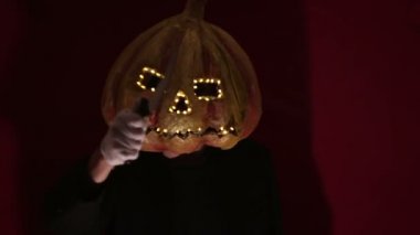 Scary man with a pumpkin head and luminous eyes looks at the camera. Man with a pumpkin head scares raising his hands up. Halloween.
