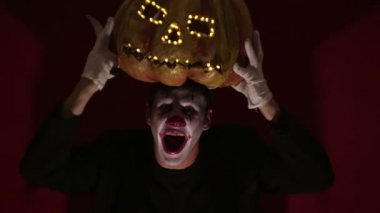 Scary man in the makeup of a clown holds a pumpkin for Halloween and puts it on his head.Terrible clown puts on a mask in the shape of a pumpkin on his head. Terrible clown holds in his hands a Jack-O