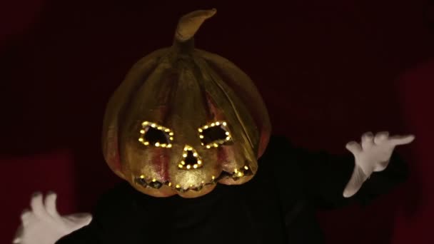 Scary man with a pumpkin head and luminous eyes looks at the camera. Man with a pumpkin head scares raising his hands up. Halloween. — Stock videók