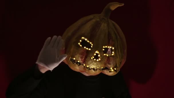 Scary man with a pumpkin head and luminous eyes looks at the camera. Man with a pumpkin head scares raising his hands up. Halloween. — Stockvideo