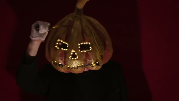 Scary man with a pumpkin head and luminous eyes looks at the camera. Man with a pumpkin head scares raising his hands up. Halloween. — Stok video