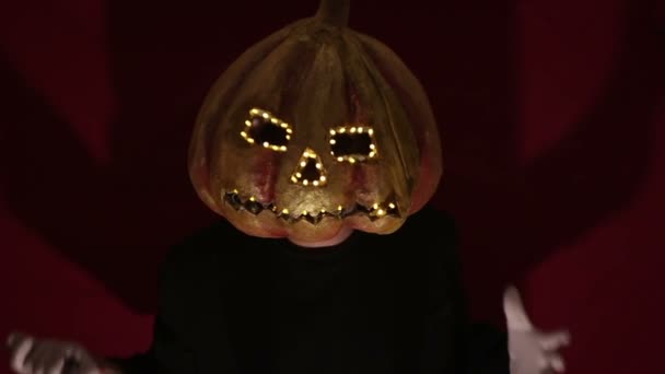 Scary man with a pumpkin head and luminous eyes looks at the camera. Man with a pumpkin head scares raising his hands up. Halloween. — 图库视频影像