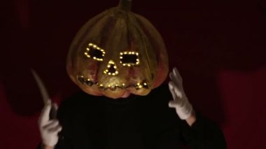 Scary man with a pumpkin head and luminous eyes looks at the camera. Man with a pumpkin head scares raising his hands up. Halloween.