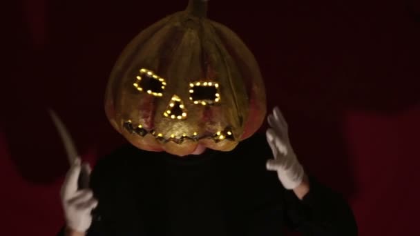 Scary man with a pumpkin head and luminous eyes looks at the camera. Man with a pumpkin head scares raising his hands up. Halloween. — Stok video
