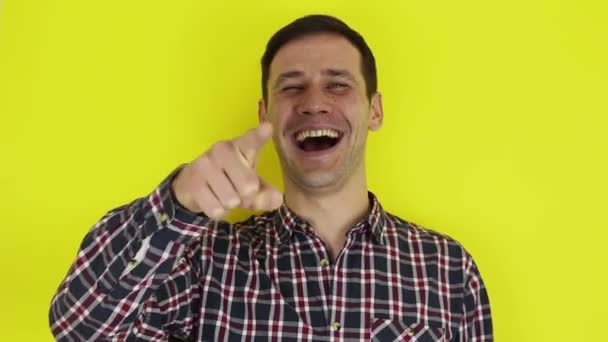 A young handsome guy in a plaid shirt looks at the camera, laughs expressively and points a finger at the interlocutor. Portrait on a yellow background. — Stock video