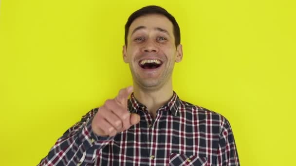 En ung flot fyr i en plaid skjorte ser på kameraet, griner udtryksfuldt og peger en finger på samtalepartner. Portræt på gul baggrund . – Stock-video