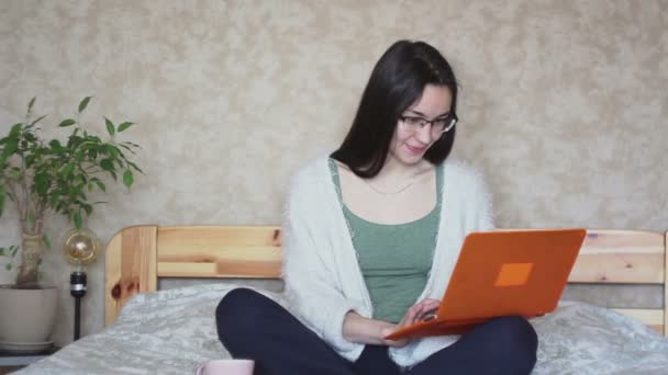 年轻迷人的女孩正坐在床上积极浏览笔记本电脑上的新闻 年轻可爱的女孩坐在床上 正在笔记本电脑上工作 在家工作 — 图库视频影像