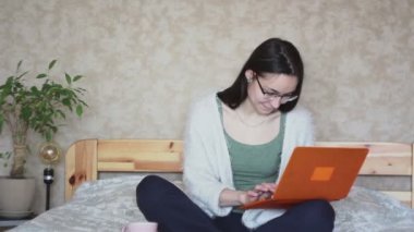 Genç çekici kız dizüstü bilgisayarıyla çalışıyor, evdeki yatakta oturuyor. Genç tatlı kız yatakta oturuyor ve dizüstü bilgisayarında yazıyor. Evde çalış..
