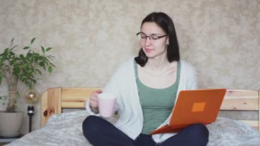 Genç ve çekici bir kız laptopuyla çalışırken pembe bir fincandan kahve içiyor, evde yatağında oturuyor. Genç ve çekici bir kız dizüstü bilgisayarıyla çalışıyor, evdeki yatakta oturuyor. Evde çalış..
