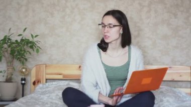 Genç ve çekici bir kız laptopuyla çalışırken pembe bir fincandan kahve içiyor, evde yatağında oturuyor. Genç ve çekici bir kız dizüstü bilgisayarıyla çalışıyor, evdeki yatakta oturuyor. Evde çalış..