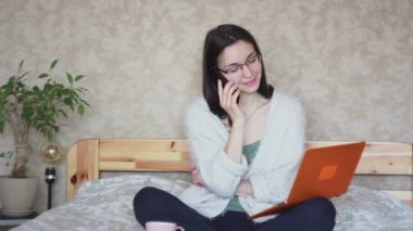 Genç ve çekici bir kız cep telefonuyla konuşuyor ve bilgisayardaki verileri karşılaştırıyor. Genç ve çekici bir kız dizüstü bilgisayarı üzerinde çalışıyor ve bir telefona cevap veriyor. Evde çalış..