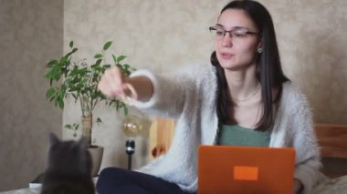 Genç çekici bir kız yatakta oturuyor ve kedisiyle oynuyor. Genç çekici bir kız evde çalışıyor, bir kediyle oynuyor. Evde çalış..