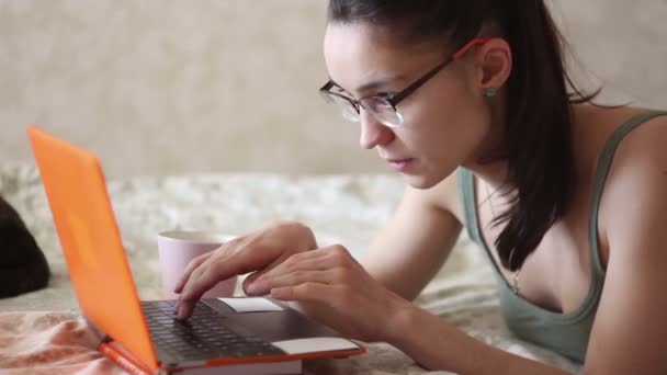 年轻漂亮的姑娘正躺在床上 在笔记本电脑上工作 一个年轻可爱的女孩躺在床上 在笔记本电脑上打字 在家工作 — 图库视频影像