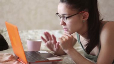 Genç çekici kız dizüstü bilgisayarıyla çalışıyor, evdeki yatakta yatıyor. Genç ve tatlı bir kız yatağında dizüstü bilgisayarıyla yazı yazıyor..