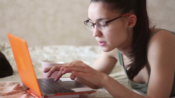 Joven chica atractiva está trabajando en su computadora portátil, acostado en la cama en casa. Una joven linda chica está acostada en la cama y escribiendo en su computadora portátil . — Vídeos de Stock