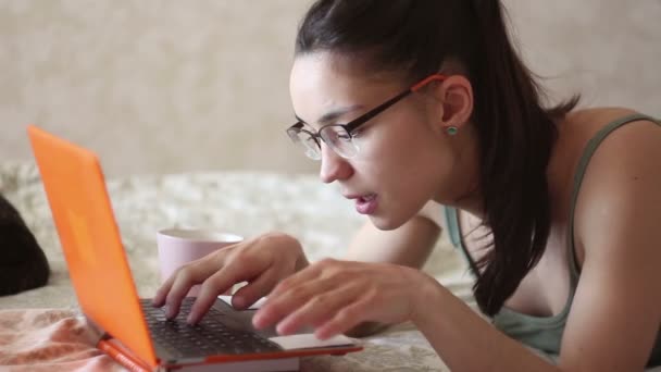 Joven chica atractiva está trabajando en su computadora portátil, acostado en la cama en casa. Una joven linda chica está acostada en la cama y escribiendo en su computadora portátil . — Vídeos de Stock