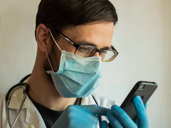 一位戴着眼镜和医疗面罩的年轻英俊医生通过电话为病人提供视频聊天咨询 一名年轻医生在检疫期间通过视频聊天为病人提供网上咨询 图库图片