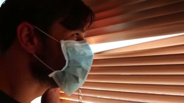 Tıbbi maske takmış genç yakışıklı bir adam pencere perdelerinden içeri bakıyor. Karantina sırasında genç bir adam pencereden bakar. Tıbbi maskeli genç bir adam pencereden bakar, kendini izole eder..