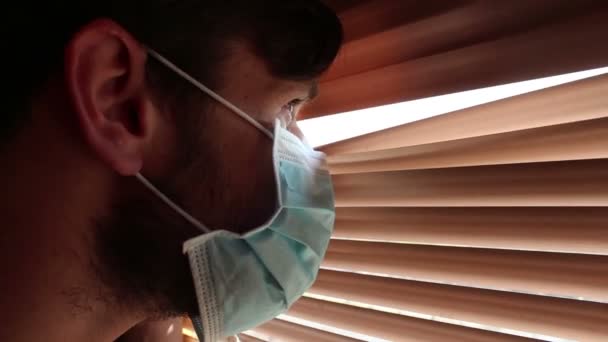 Tıbbi Maske Takmış Genç Yakışıklı Bir Adam Pencere Perdelerinden Içeri — Stok video