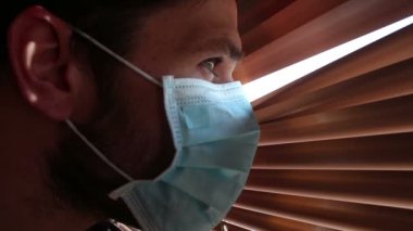 Tıbbi maske takmış genç yakışıklı bir adam pencere perdelerinden içeri bakıyor. Tıbbi maskeli genç bir adam pencereden bakar, kendini izole eder..
