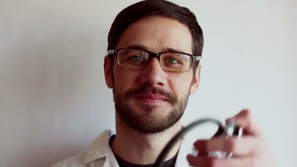 眼鏡をかけた若いハンサムな医者が患者の診察の準備をしながら聴診器を装着する 若い医者が聴診器を持ってる — ストック動画