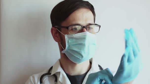 患者の診察の準備をしながら眼鏡と医療用マスクの若いハンサムな医者が滅菌手袋を装着します 若い医者が患者の診察の準備をしている 保護下の医者は — ストック動画