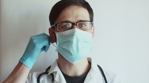 眼鏡をかけた若いハンサムな医者は 彼の医療マスクを脱いで安心してため息をつく 若い医者は患者の回復を喜び 彼の保護マスクを脱いで安心してため息をつく — ストック動画