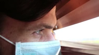 Tıbbi maske takmış genç yakışıklı bir adam pencere perdelerinden içeri bakıyor. Tıbbi maskeli genç bir adam pencereden bakar, kendini izole eder..